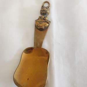 May include: A vintage brass spoon with a Cheshire Cat design. The spoon features a scoop-shaped bowl and a handle with a cat's head and the words "Cheshire Cat". The spoon is a warm gold color.