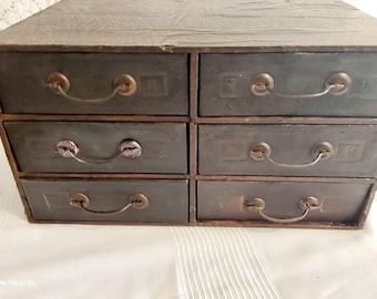 May include: A vintage six-drawer chest in dark grey with antique brass handles. The drawers are arranged in two columns of three, with a slightly distressed look. The chest is likely made of wood and is suitable for storing small items.