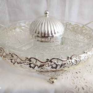 Puede incluir: Una fuente de vidrio transparente con base, asas y tapa de metal plateado. La fuente tiene un patrón floral decorativo alrededor del borde y está dividida en secciones. Las asas son curvas y ornamentadas.