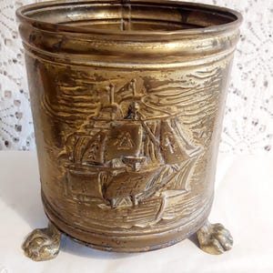 Könnte beinhalten: Ein antiker Messingbehälter mit detailliertem Relief eines Segelschiffs. Der zylindrische Behälter steht auf drei Klauenfüßen und hat einen breiten Rand. Das Messing hat eine warme, gealterte Patina. Die Höhe beträgt etwa 15 cm.