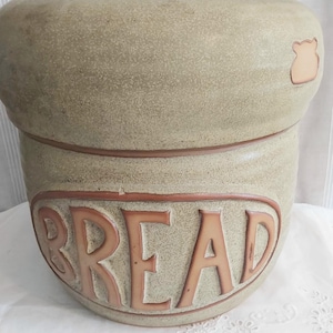 May include: A beige ceramic bread crock with a speckled texture and a thick, rounded rim. The word BREAD is embossed in large, capital letters across the front in a reddish-brown colour. A small, stylised bear-shaped icon is visible on the upper right.