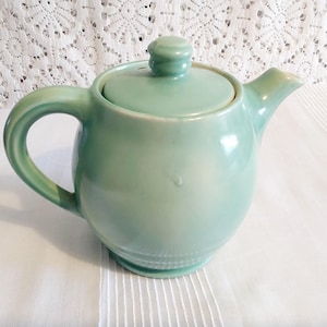 May include: A light green ceramic teapot with a lid, handle, and spout. The teapot has a rounded body and a small knob on the lid. The handle is curved, and the spout is short and angled. The teapot is sitting on a white surface.