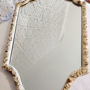 Puede incluir: Espejo ornamentado de estilo antiguo, con marco crema y detalles dorados. El espejo tiene una forma curva única y probablemente es de vidrio. El marco presenta diseños florales y de volutas detallados, lo que añade a su estética vintage.