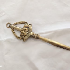 Könnte beinhalten: Ein Vintage-Brieföffner aus Messing mit dekorativem Griff. Der Griff trägt den Text "LIGHTHOUSE LANDS END" und ein detailliertes Relief eines Schiffes. Der Öffner hat eine hellgoldene Farbe und ist vor einem weißen Hintergrund platziert.