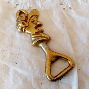 May include: A vintage brass bottle opener with a whimsical design. The opener features a stylized face with a hat and a loop for hanging. The brass has a golden hue with some signs of wear, adding to its antique charm.