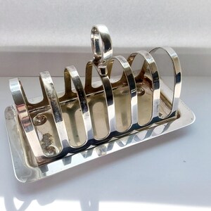 May include: A silver-colored toast rack with six arched slots and a handle. The rack sits on a rectangular base. The design is simple and elegant, suitable for serving toast at the breakfast table.