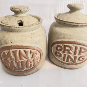 Puede incluir: Dos tarros de almacenamiento de cerámica beige moteada con tapas a juego. Un tarro presenta un emblema ovalado marrón con el texto en relieve MINT SAUCE y una muesca en la tapa. El segundo tarro tiene un emblema marrón con el texto DRIPPING.