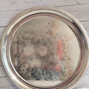 May include: A round silver-coloured metal serving tray featuring an intricate embossed floral and scrollwork pattern across its surface. The tray has a raised, textured rim with a repeating vertical line design along the outer edge of the border.