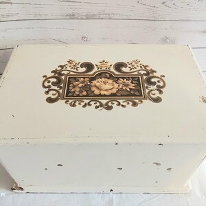 May include: A rectangular off-white storage box with a hinged lid. The top surface features a central rectangular panel containing a floral illustration of roses and leaves, surrounded by decorative brown scrollwork and ornate filigree patterns.