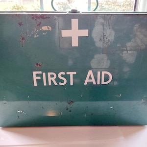 May include: A teal-coloured vintage first aid box, featuring a white cross and the words "FIRST AID" in white. The metal box shows signs of wear and tear. A handle is visible at the top. The box is likely made of metal.
