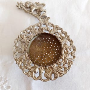 Puede incluir: Infusor de té plateado adornado con una figura de león decorativa y un intrincado diseño floral. El infusor tiene una cesta redonda perforada para hojas de té. Un accesorio de cocina de estilo vintage.