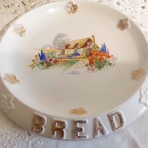 May include: White ceramic bread plate with a colorful cottage scene. The rim features embossed flowers and gold accents. The word "BREAD" is embossed in gold lettering at the bottom of the plate. A decorative piece for the kitchen.