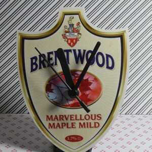 May include: A shield-shaped clock with a gold border and a white background. The clock face features a red maple leaf design and the text "Brentwood Marvellous Maple Mild 3.7% alc. vol."