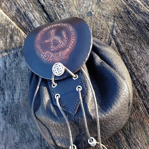 May include: A black leather bag with a flap featuring a brown embossed design of a cat and the text "TOUCH NOT THE CAT BUT A GLOVE". The bag has a drawstring closure with silver metal beads.