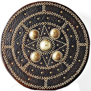 May include: A round, dark brown leather shield with intricate Celtic knotwork designs and gold metal accents. The shield is decorated with numerous gold studs and five large, round, gold metal bosses.
