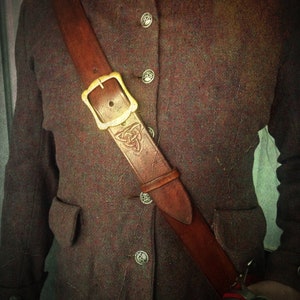 Könnte beinhalten: Ein braunes Lederband mit einer Messing-Schnalle und einem dekorativen Design. Das Band wird über einer braunen Tweedjacke getragen.