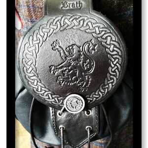 Puede incluir: Sporran de cuero negro con un diseño de nudo celta y un emblema de león. El sporran tiene un botón plateado con un diseño de cardo y una correa con el texto "Clan Braith".