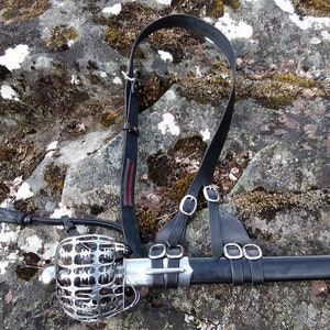 May include: A black leather sword scabbard with a metal guard and a black leather strap. The scabbard is hanging from a strap with two buckles.