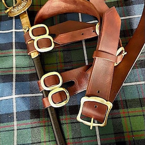 May include: A brown leather sword belt with gold buckles and a matching sword scabbard. The belt and scabbard are laid out on a green and blue plaid fabric.