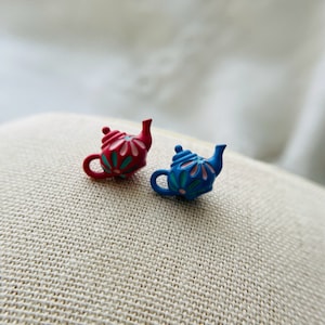 Może przedstawiać: Dwa miniaturowe czajniczki, jeden czerwony z różowymi kwiatowymi detalami, a drugi niebieski z turkusowymi kwiatowymi detalami, na beżowym tle z tkaniny.