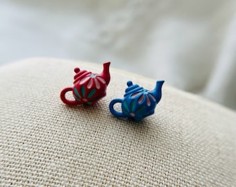 May include: Two miniature teapots, one red with pink floral details and one blue with turquoise floral details, on a beige fabric background.