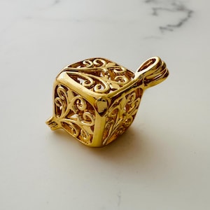 Può includere: Un ciondolo dorato, ornato, con lavorazione traforata a forma di cubo con un anello in alto per attaccarlo a una catena.