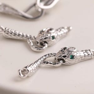 Peut inclure: Gros plan sur plusieurs apprêts de bijoux argentés en forme de serpent. Chaque tête de serpent a un petit œil en pierre précieuse verte. Les apprêts sont détaillés avec des écailles texturées et une queue incurvée.