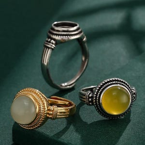 Könnte beinhalten: Drei Ringe mit aufwendigen Designs werden auf einer dunkelgrünen Oberfläche gezeigt. Ein Ring ist silberfarben mit einer detaillierten Fassung. Ein weiterer Ring ist goldfarben mit einem hellfarbigen Stein. Der dritte Ring ist silberfarben mit einem gelben Stein.
