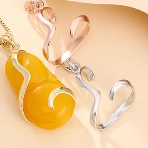 Op de afbeelding: Een close-up van een gele, kalebasvormige hanger met een goudkleurig frame, hangend aan een gouden ketting. Twee decoratieve metalen stukken, één roségoud en één zilver, zijn ook zichtbaar, tegen een lichtbeige achtergrond.