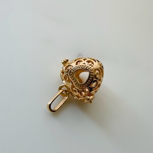 May include: Gold heart-shaped locket with intricate filigree detailing. The locket has a small opening at the top for adding a chain.