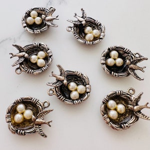 May include: Ten silver charm pendants shaped like bird nests with three white pearl-like eggs and a small silver bird.