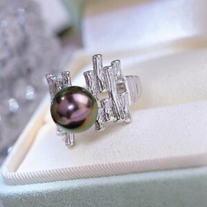 May include: A silver ring featuring a large, iridescent dark pearl. The ring has a unique, textured design with a branch-like pattern around the pearl. The ring is set against a soft, light background.
