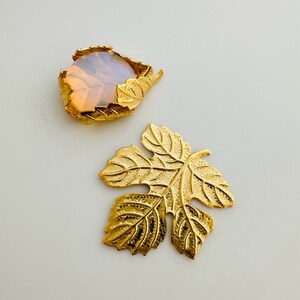 May include: Two gold-coloured leaf-shaped jewellery components. One features a round, opalescent stone set within a gold leaf frame. The other is a solid gold leaf with detailed veining, ideal for crafting or jewellery making.