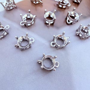 May include: A collection of silver-coloured metal jewellery settings, each featuring a circular base with four prongs extending upwards to hold a stone. Each setting includes a small loop on the side for attachment, designed for crafting necklaces or earrings.