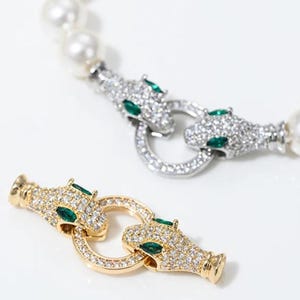 May include: Two jewellery clasps featuring dual panther heads with clear pavé stones and green marquise-cut eyes, one in silver tone and one in gold tone. The silver clasp is attached to a white pearl strand, while the gold clasp is separate.
