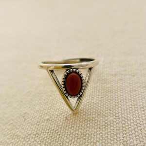 Può includere: Un anello d'argento con un design triangolare e una pietra ovale rossa al centro.