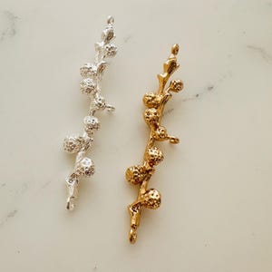 Könnte beinhalten: Zwei Metall-Anhänger in Form eines Zweigs mit kleinen runden Beeren. Der Anhänger links ist silberfarben und der Anhänger rechts ist goldfarben.