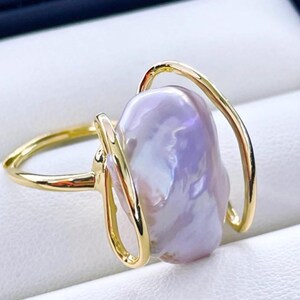 May include: A gold-colored ring featuring a large, irregularly shaped, lavender-hued pearl. The pearl is held in place by a delicate, openwork design. The ring's band and the pearl's setting have a polished, reflective surface.