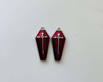 Op de afbeelding: Twee zilverkleurige, kistvormige bedels met een rode emaille afwerking en een zilveren kruisontwerp.