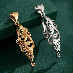 May include: Two ornate pendants, one gold and one silver, are displayed on a dark green surface. Each pendant has a detailed, swirling filigree design and a loop for a chain. These pendants are likely for necklaces.