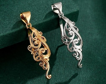 May include: Two ornate pendants, one gold-toned and one silver-toned, rest on a dark green surface. Each pendant features a detailed, swirling filigree design with a loop at the top for a chain. The pendants are likely for necklaces.