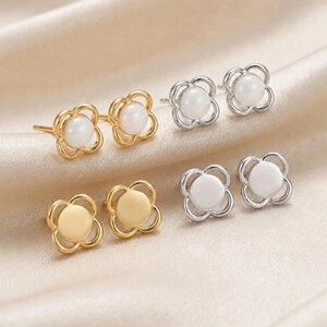 May include: Four pairs of stud earrings in gold and silver tones. Each earring features a four-petaled flower frame surrounding a central circular element. Two pairs have white pearl-like centers, while the other two have solid metal centers.