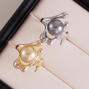Può includere: Due anelli con design organici e fluidi. Uno presenta una montatura color oro con una perla color crema. L'altro anello ha una montatura color argento con una perla grigia. Entrambi gli anelli sono su una superficie beige chiaro.