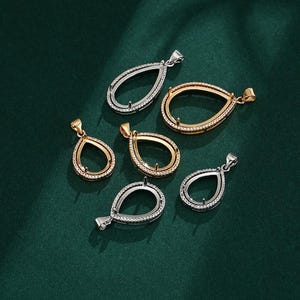 May include: An assortment of teardrop-shaped pendant settings in gold and silver tones. Some settings are encrusted with small, clear stones. The pendants are displayed on a dark green surface, showcasing their intricate designs and potential for jewelry making.