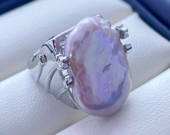 May include: A close-up of a silver ring featuring a large, iridescent, baroque pearl. The pearl has a soft, lavender hue with subtle blue and pink highlights. The ring's band has a detailed, ornate design with small, decorative accents.