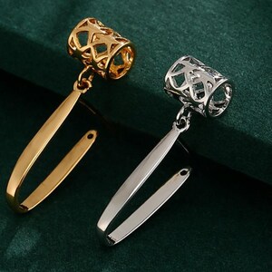 May include: Two metal hair cuffs, one gold and one silver, are displayed on a dark green surface. Each cuff has an openwork cylinder and a curved, elongated bar. The gold cuff has a shiny finish, while the silver cuff has a polished appearance.
