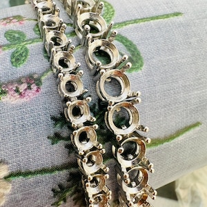 May include: Two silver-colored metal bracelet settings with round bezel cups and prongs. The settings are arranged in parallel rows, showcasing the design for jewelry making. The background features a floral pattern on a light blue fabric.
