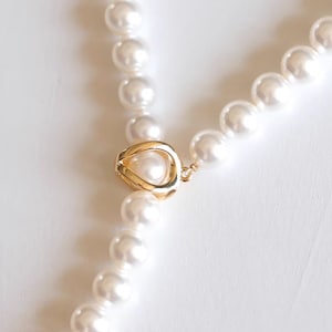 May include: A white pearl necklace with a gold clasp. The clasp is shaped like a teardrop with a circle in the center.