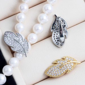 Può includere: Primo piano di una collana di perle con una chiusura a forma di piuma argentata e una dorata. Le chiusure sono incastonate con piccoli cristalli scintillanti. La collana è esposta su una superficie color crema.