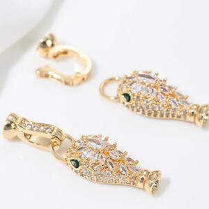 May include: Two gold-toned metal fish-shaped jewellery charms encrusted with clear marquise-cut cubic zirconia stones and small green stone eyes, each featuring a gold-toned metal bail attachment for connecting to a chain or necklace cord.